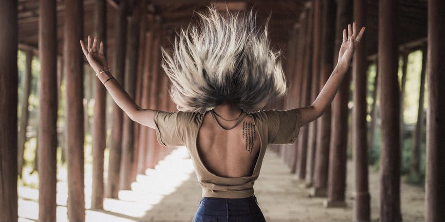 An Asian woman in a tunnel wearing a backless t-shirt has her back to the camera and throws her white hair into the air, with her arms extended outward.