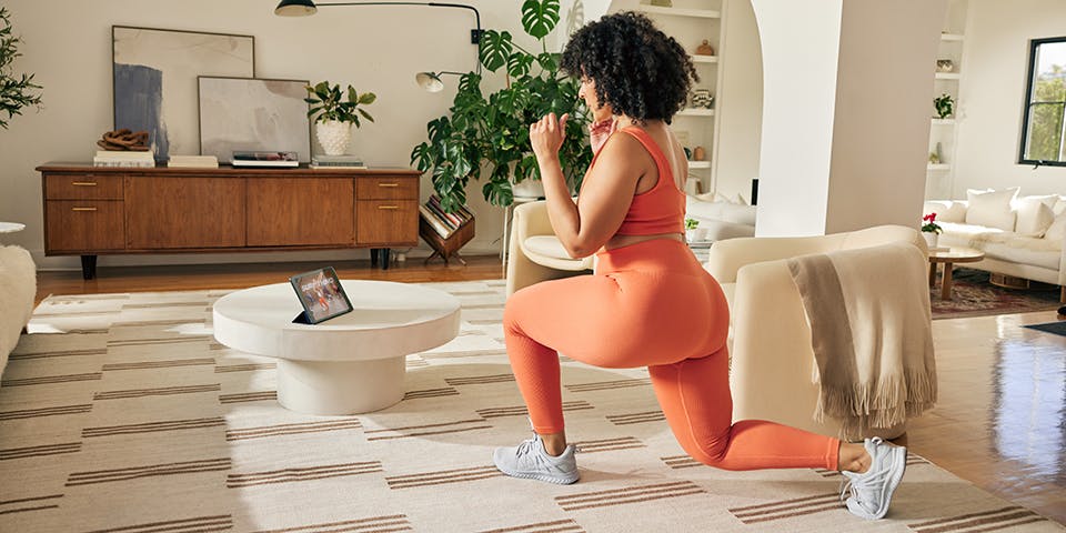 Fotografía en color de una mujer negra con curvas, con un afro vestido con ropa deportiva naranja, haciendo una zancada profunda en su sala de estar mientras ve un programa de acondicionamiento físico de BoDi en un iPad colocado sobre su mesa de café.
