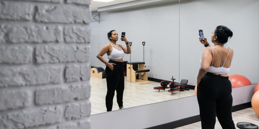 A color photo of a curvy woman with brown skin looking into a mirror at a gym, holding up her phone and taking a picture.